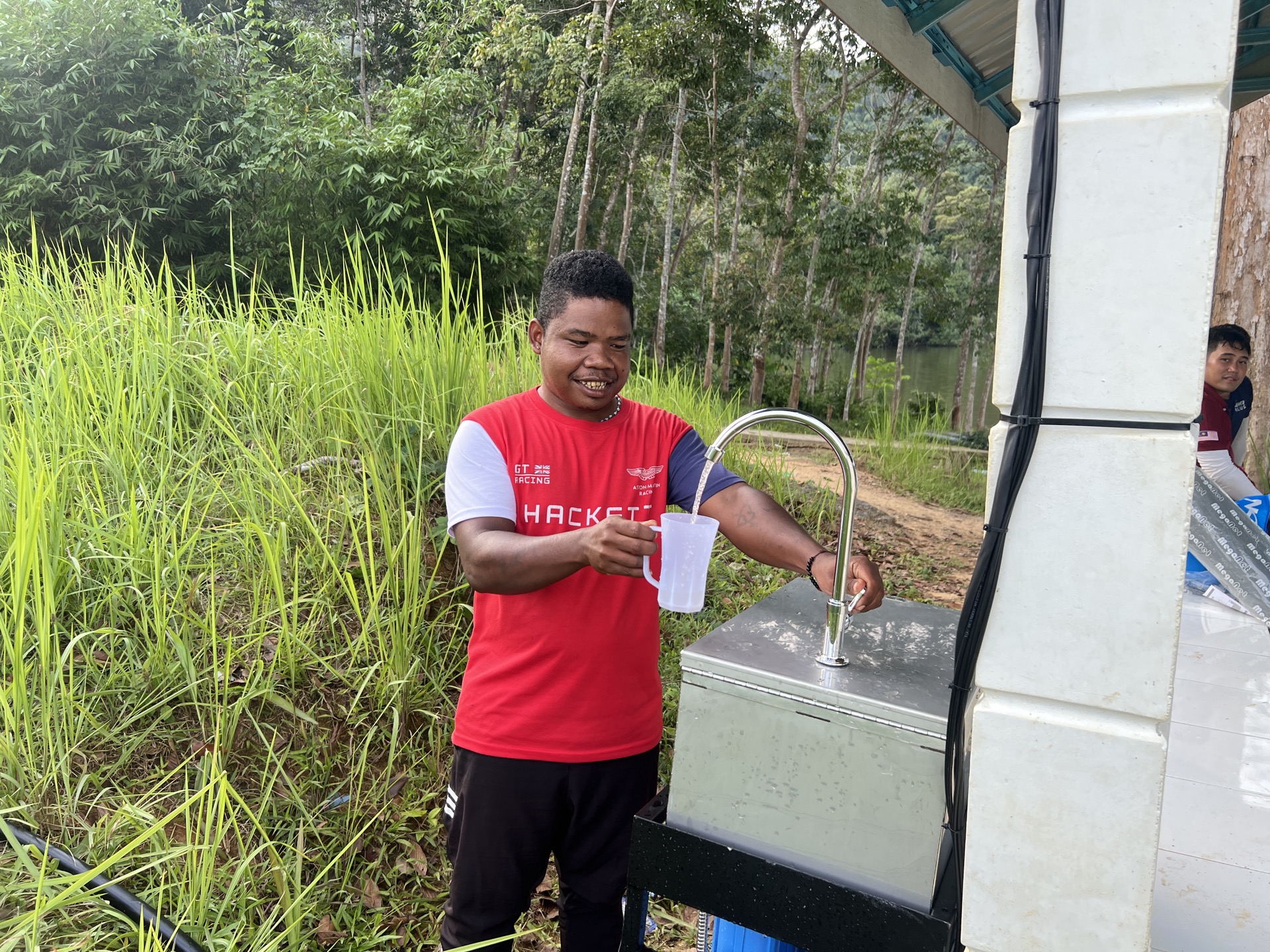 Saora+ proprietary gravity-fed water filtration system installed at the edge of an Orang Asli village, with PVC pipework connecting to a forest stream